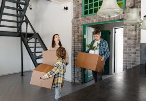 A family moving into their new home, carrying boxes and belongings indoors.